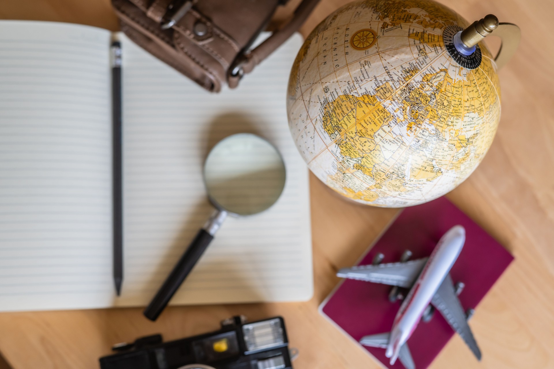 Traveler is planning a trip around the world using a passport, globe, magnifying glass, notebook and pencil, along with a small airplane model and vintage camera, on a wooden table