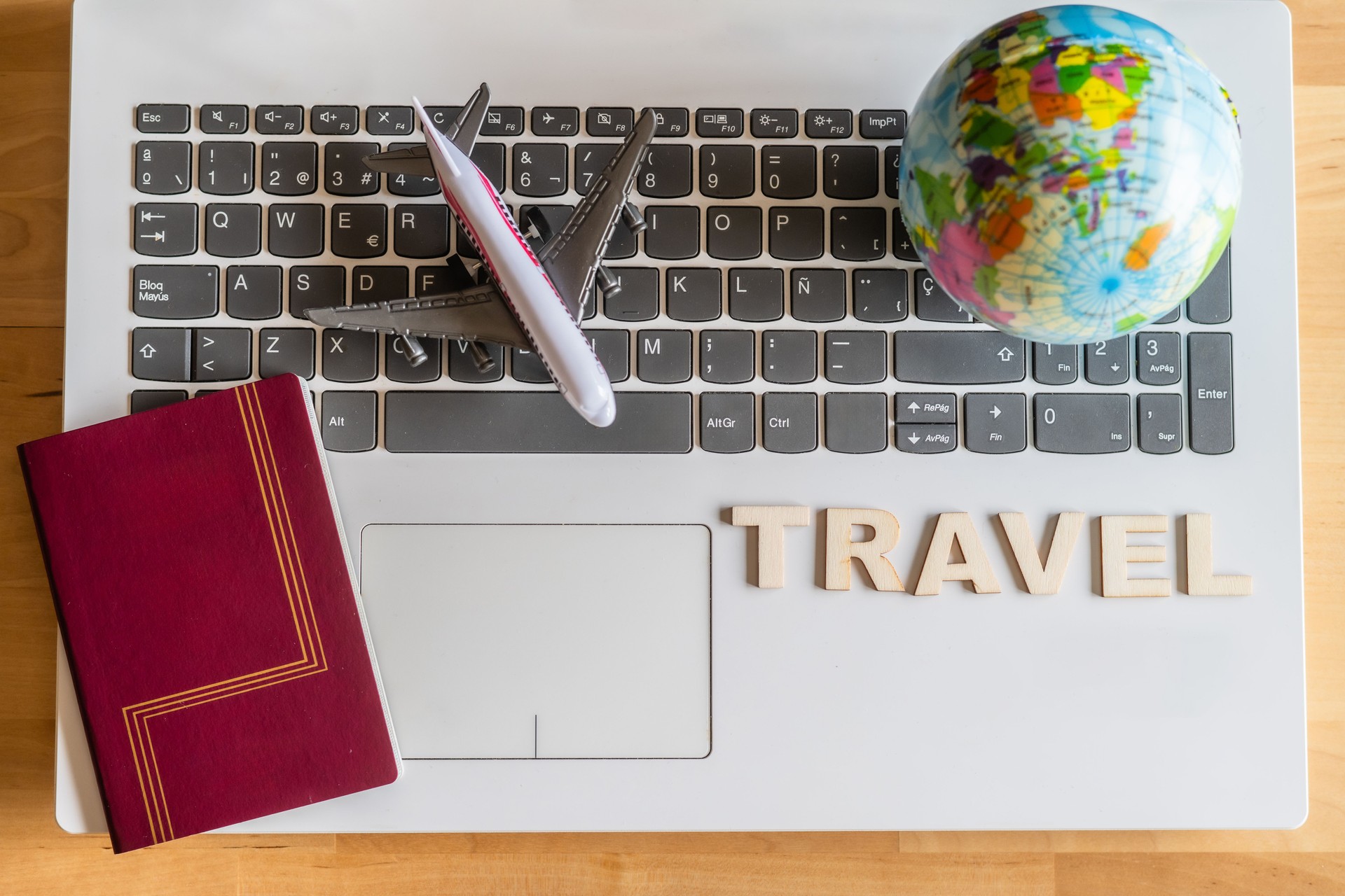 Airplane models, a world globe, a passport, and the word travel made of wooden letters are sitting on a laptop keyboard, suggesting online trip planning and booking
